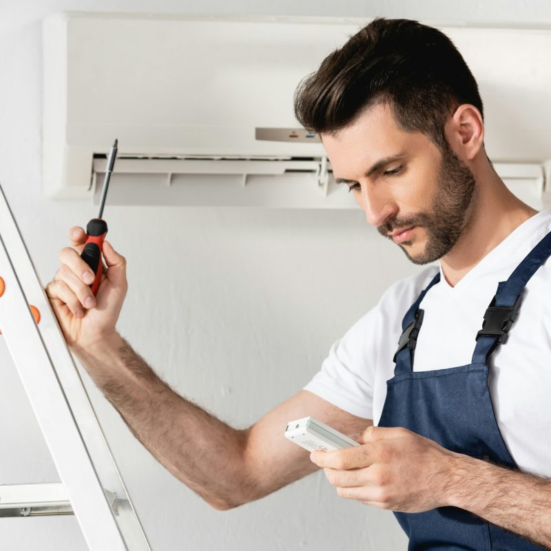 handsome-repairman-holding-remote-controller-and-screwdriver-while-standing-on-stepladder-near-air